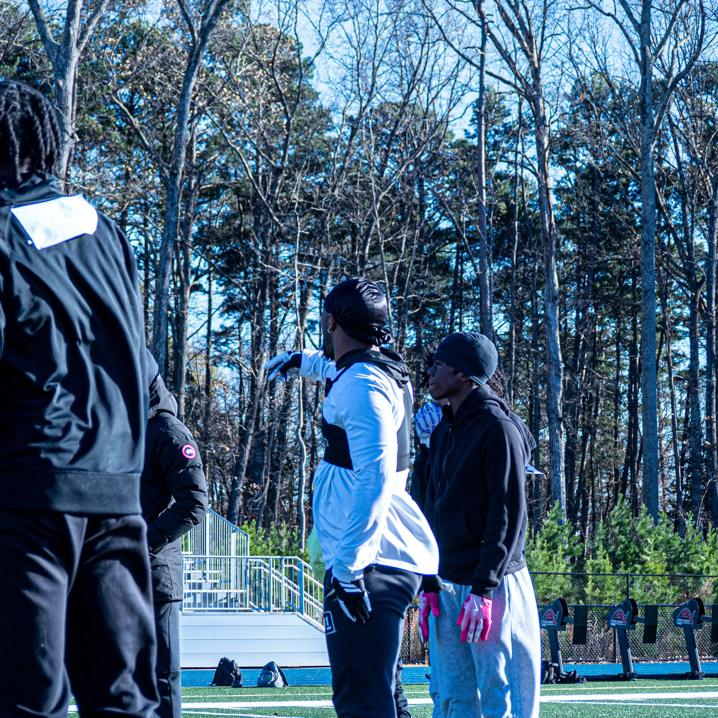 People standing on a sports field, possibly during a practice or game, with trees and a blue sky in the background.