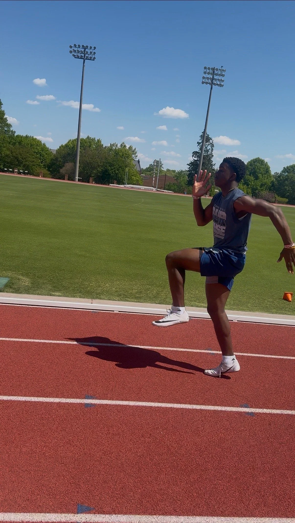 A male athlete in a gray t-shirt and blue shorts sprinting on a track, raising his left hand near his face, with a green field, track lighting poles, and a blue sky with clouds in the background.