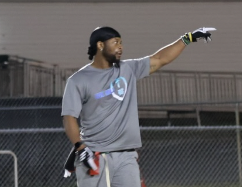 An African American man wearing a gray athletic shirt and gray shorts, standing on a sports field or track, pointing with his right arm.