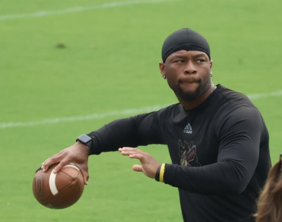 A man wearing a black sports long sleeve shirt, black head cap, and a fitness tracker on his left wrist, holding a football on a grassy field, preparing to throw it.