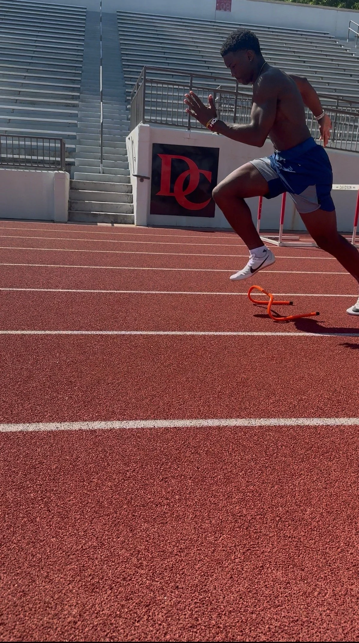 Young male athlete practicing hurdle running on a track, mid-air over a hurdle, wearing athletic shorts and sneakers.