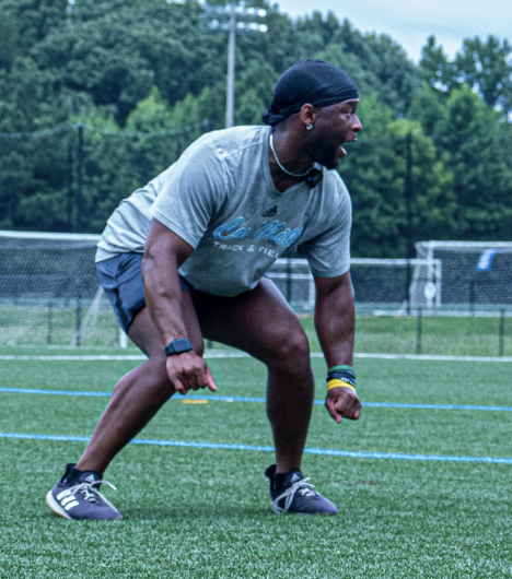 A woman in athletic gear crouching on a track field, preparing to sprint, with trees and sports facilities in the background.