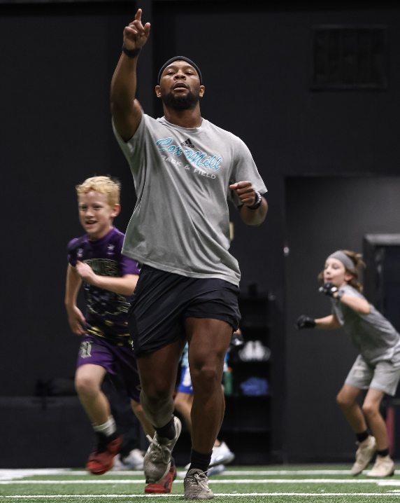 A man running indoors with children in sports attire around him, including a boy in purple and a girl in gray, during a sports training session.