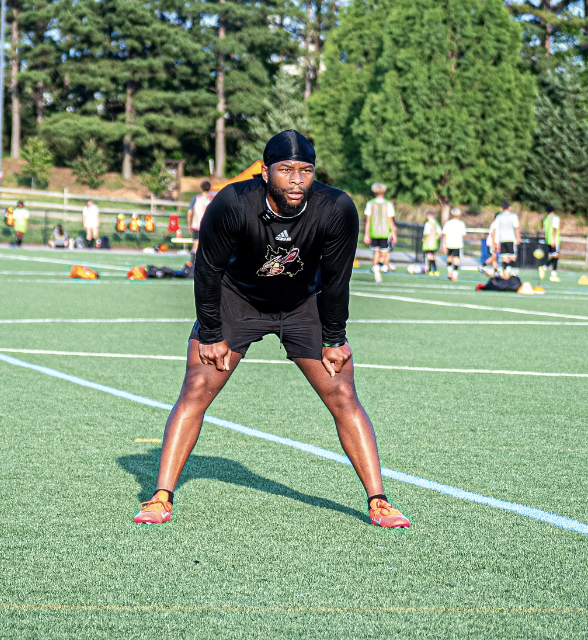 African American male athlete in black sportswear stretching on a football field with others practicing in the background.