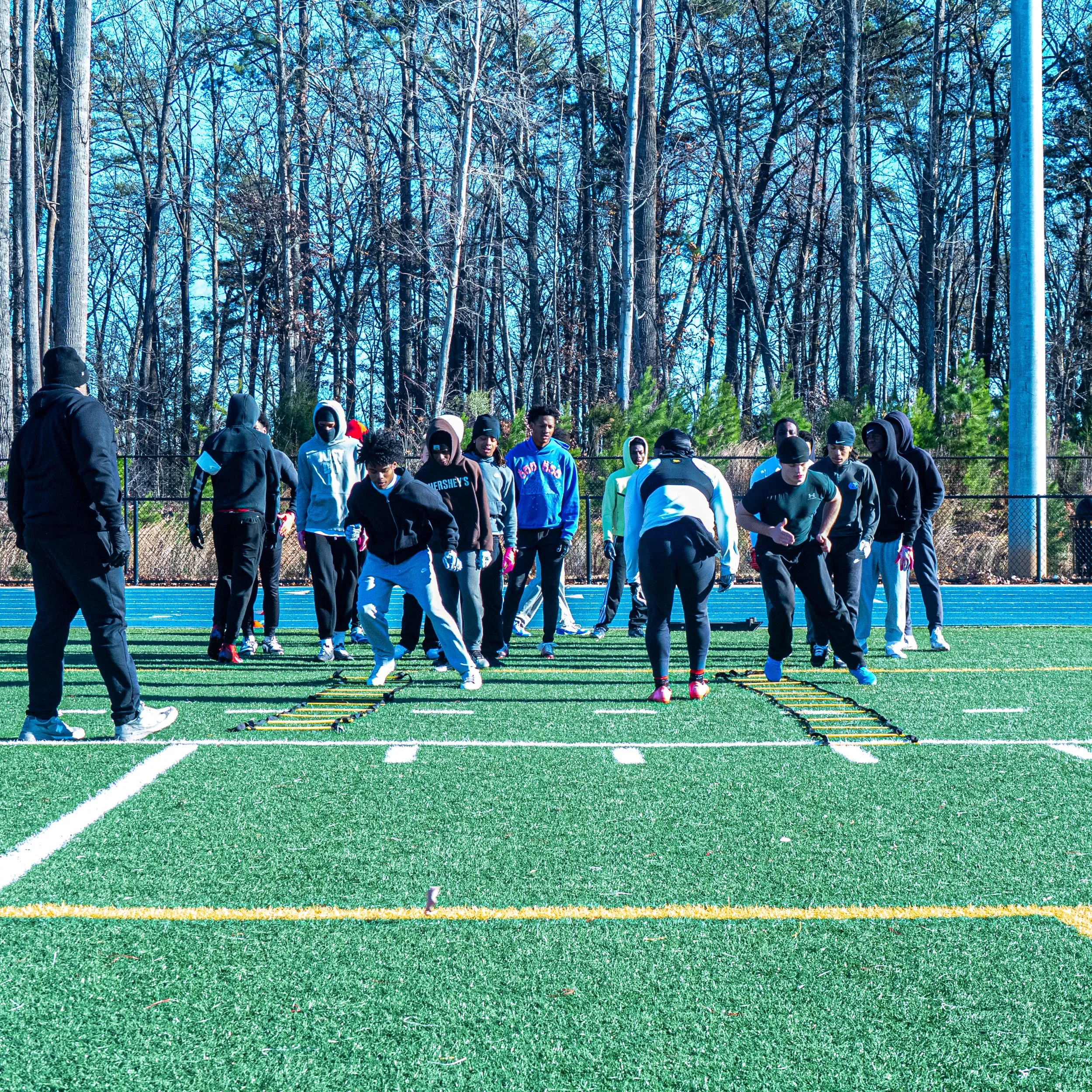 Group of young football players practicing agility drills on a football field with trees and a fence in the background.