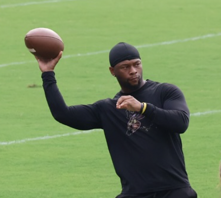 An athlete in black athletic wear and a black head covering preparing to throw a football on a grassy field.