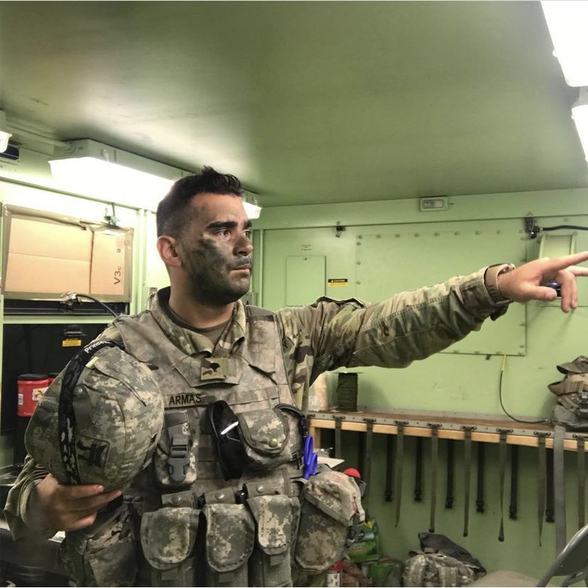 A soldier in camouflage uniform with face paint, holding a helmet, pointing to the right inside a military room.