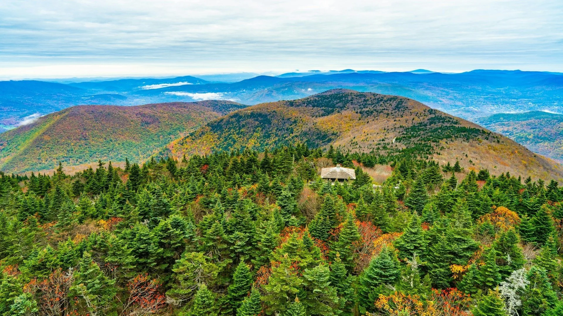 View of a mountain landscape with green forests, colorful fall foliage, and a small cabin nestled among the trees, under a cloudy sky.
