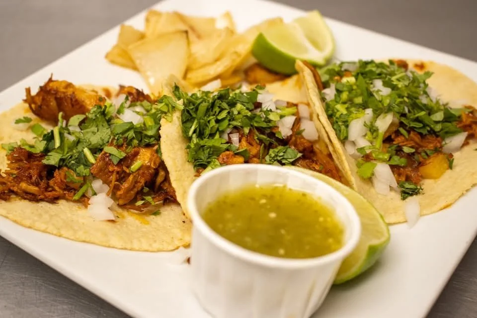 Three tacos filled with shredded meat, topped with chopped onions and cilantro, served with lime wedges, along with potato chips and a small cup of green salsa on a white plate.