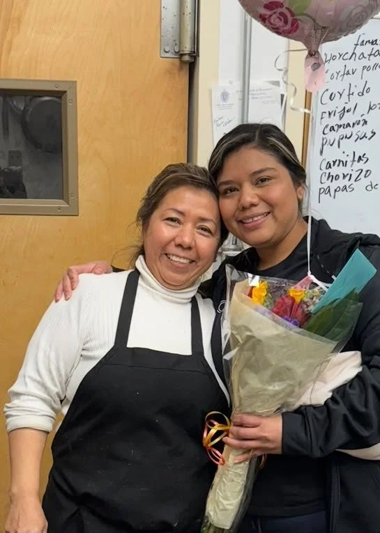 Two women smiling and hugging in an indoor setting, one holding a bouquet of flowers with a balloon above them.