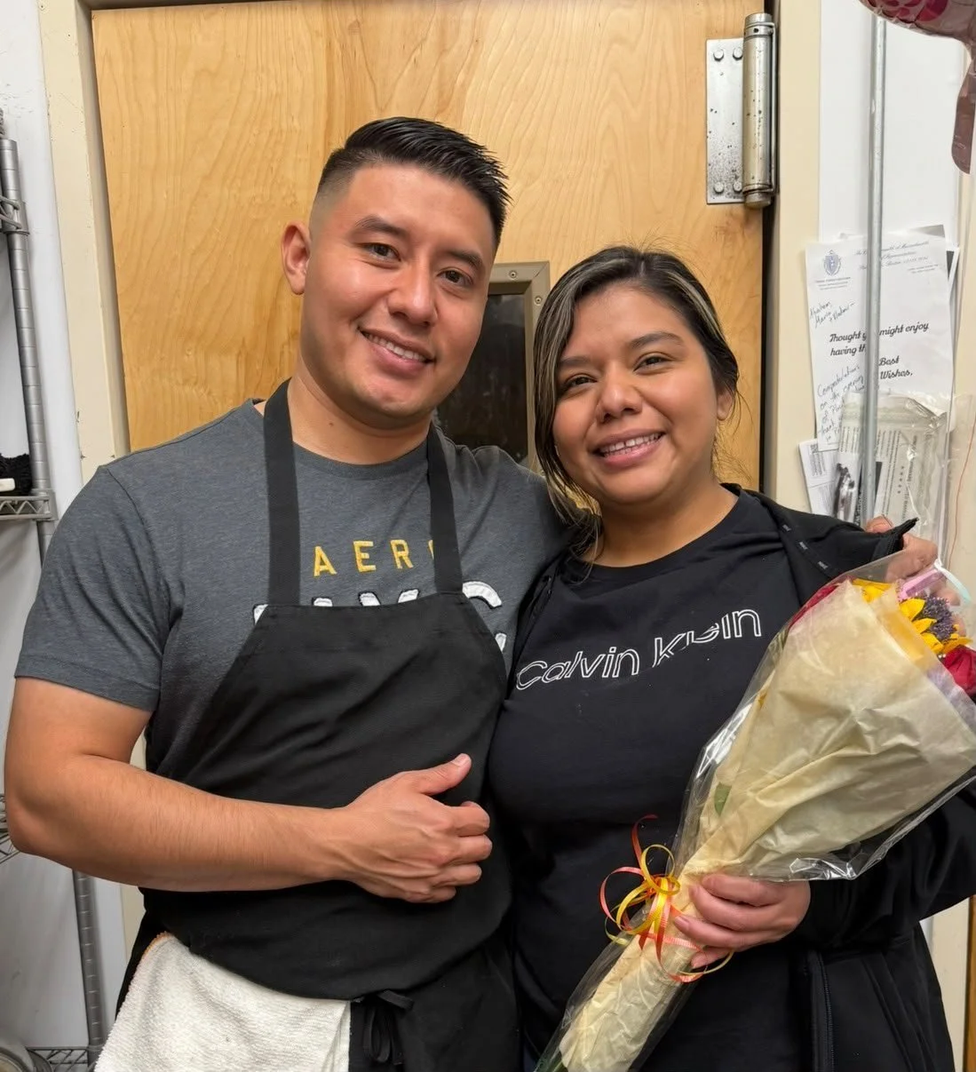 A man and woman smiling together, holding a bouquet of flowers, in a kitchen or storage room with a wooden door in the background.