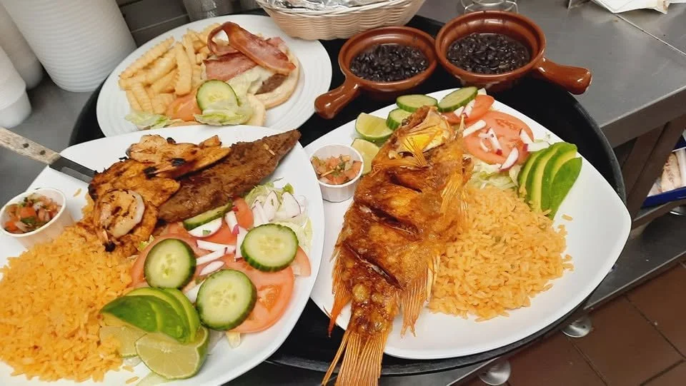Two plates of Mexican seafood dishes with cooked rice, grilled fish, and fried fish topped with sauce, accompanied by fresh lime, cucumber, tomato, and onion slices, with side sauces and a small plate of fries with bacon in the background.