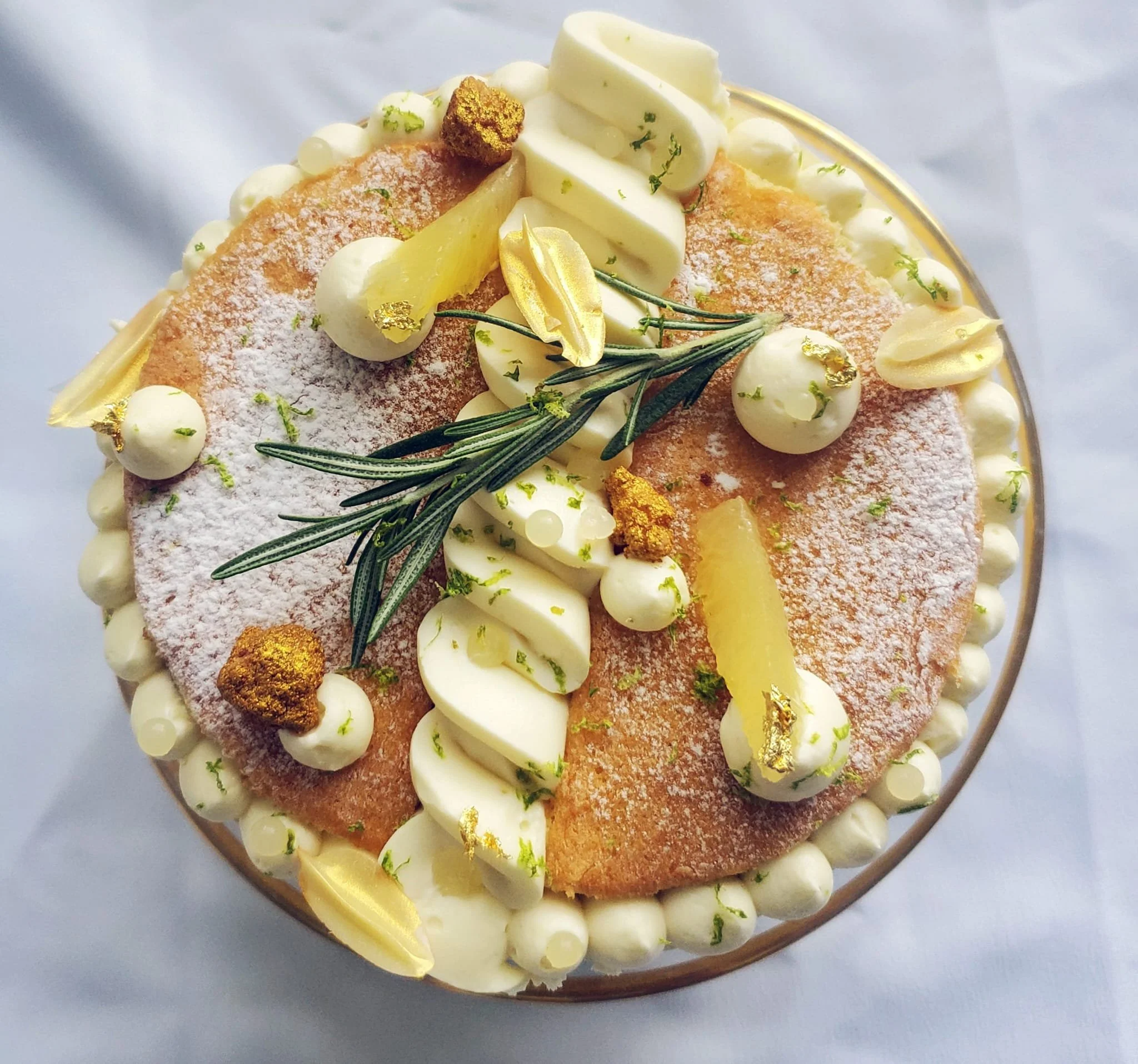 A decorated cake topped with white chocolate, gold accents, sprigs of rosemary, and powdered sugar, with a pinkish-brown frosting surface. Lemon white chocolate Homemade handcrafted French Craftmanship