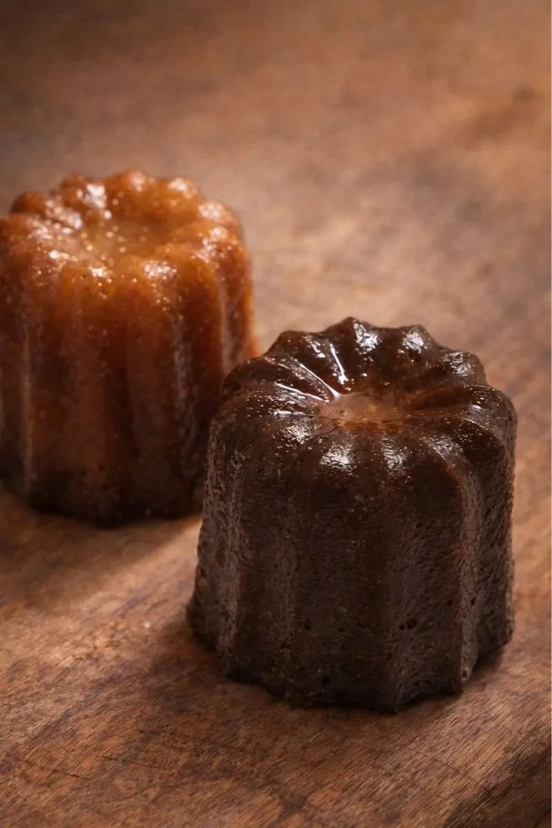 Two canelé pastries, one light brown and one dark brown, placed on a wooden surface. Homemade handcrafted French Craftmanship