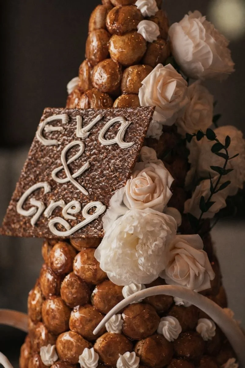 Close-up of a croquembouche with a chocolate plaque reading 'Eva & Greg' surrounded by white roses, decorated with caramel-glazed cream puffs and icing.