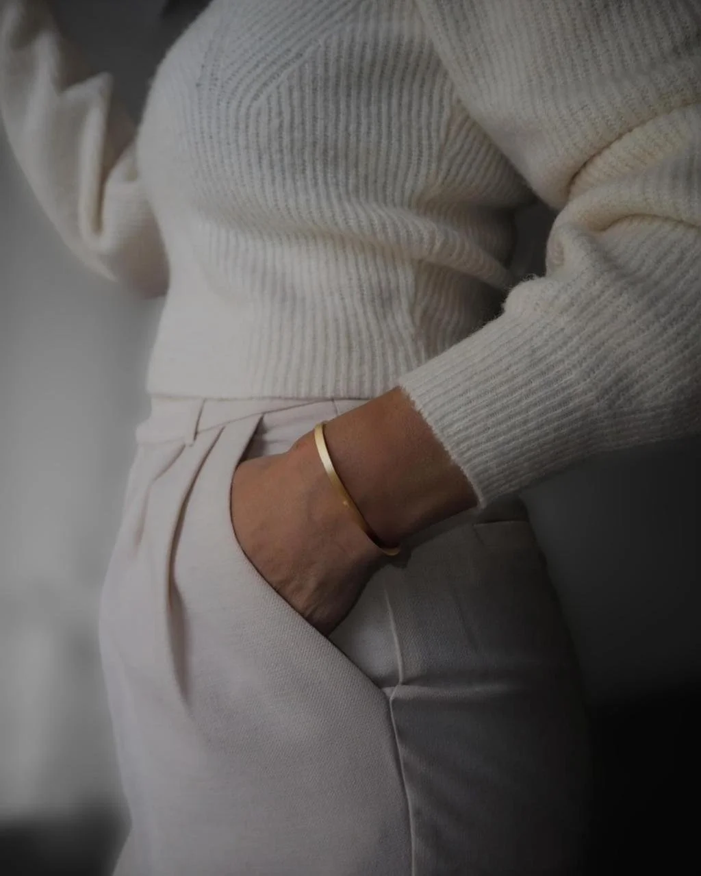Person wearing a beige knit sweater and pants, sitting with one hand in their pocket, gold bracelet on wrist, against a neutral background.