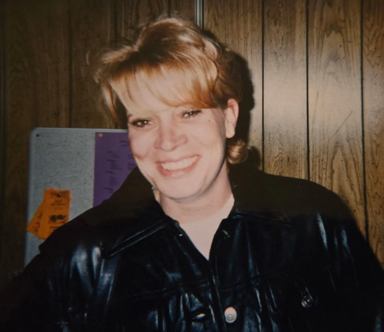 A smiling woman with short, light brown hair, wearing a black leather jacket, standing in front of a wooden wall.