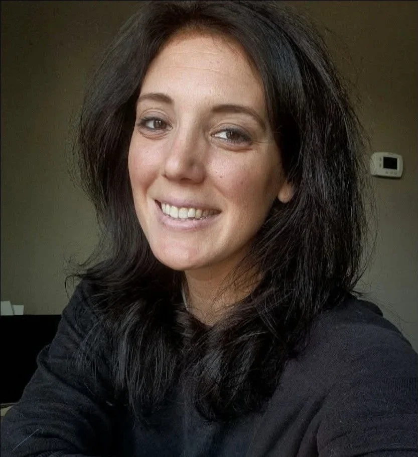 A woman with shoulder-length dark hair, smiling and wearing a black top, sitting indoors with a thermostat on the wall in the background.