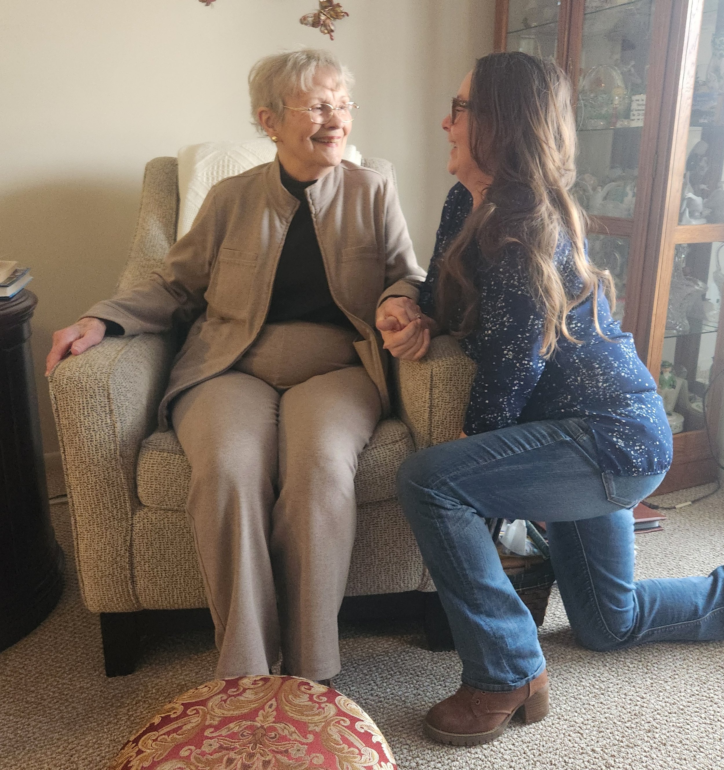 An elderly woman sitting in a beige armchair, smiling, as a younger woman kneels beside her, holding her hand. They appear to be enjoying a joyful moment together in a cozy living room.