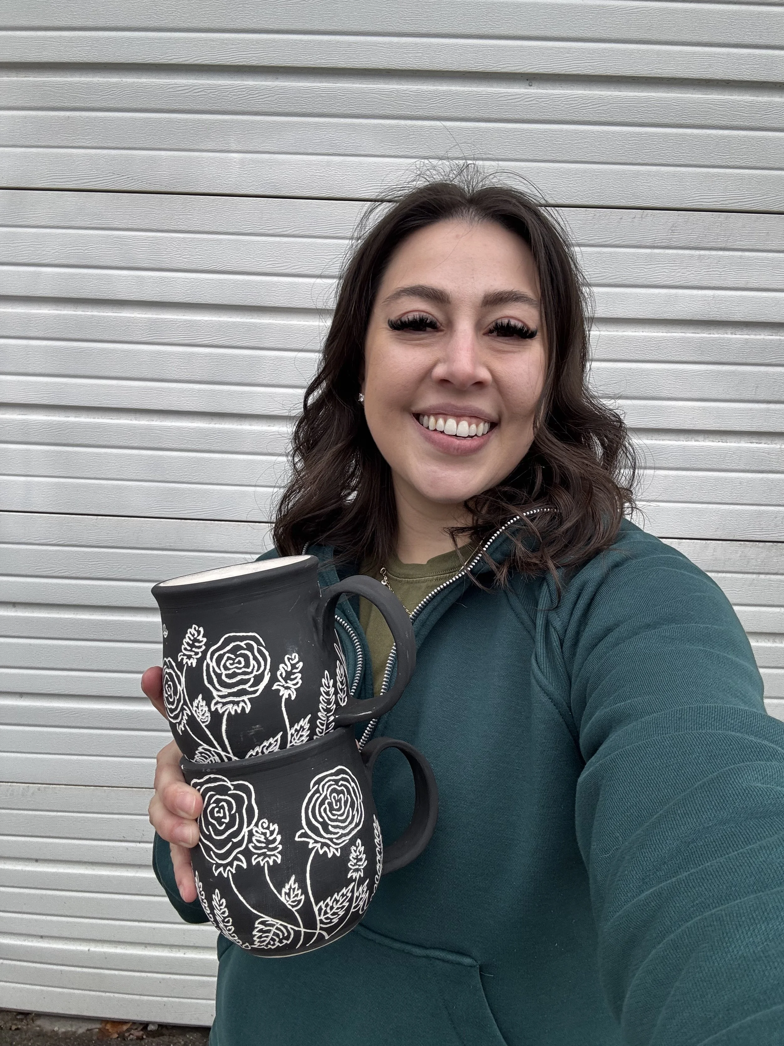 A woman with dark hair and a green jacket smiling while holding two black mugs decorated with white rose patterns in front of a gray, metal garage door.