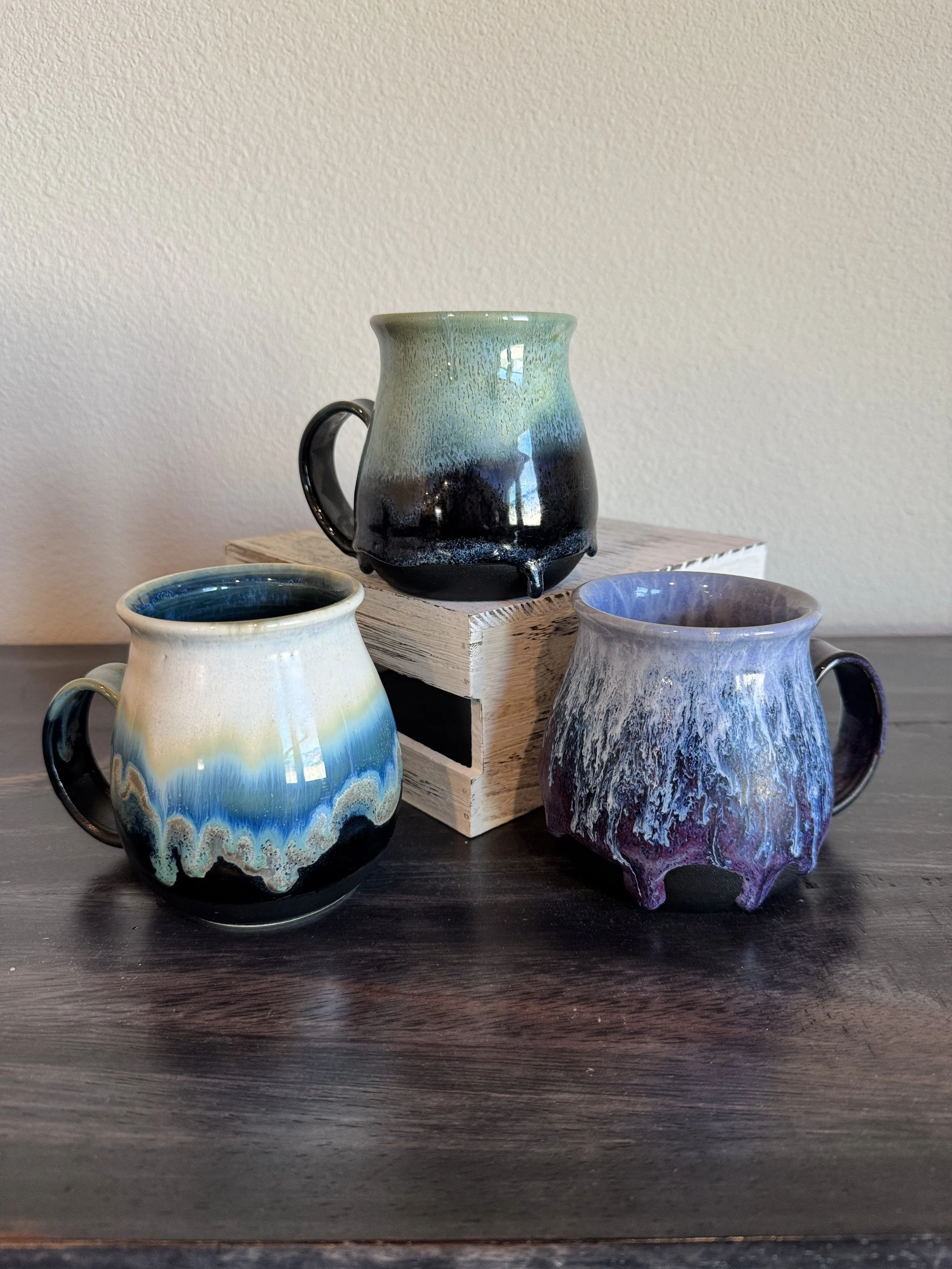 Three colorful glazed ceramic mugs with unique designs and a black handle, placed on a dark wooden table with a small wooden crate behind them and a light-colored textured wall in the background.