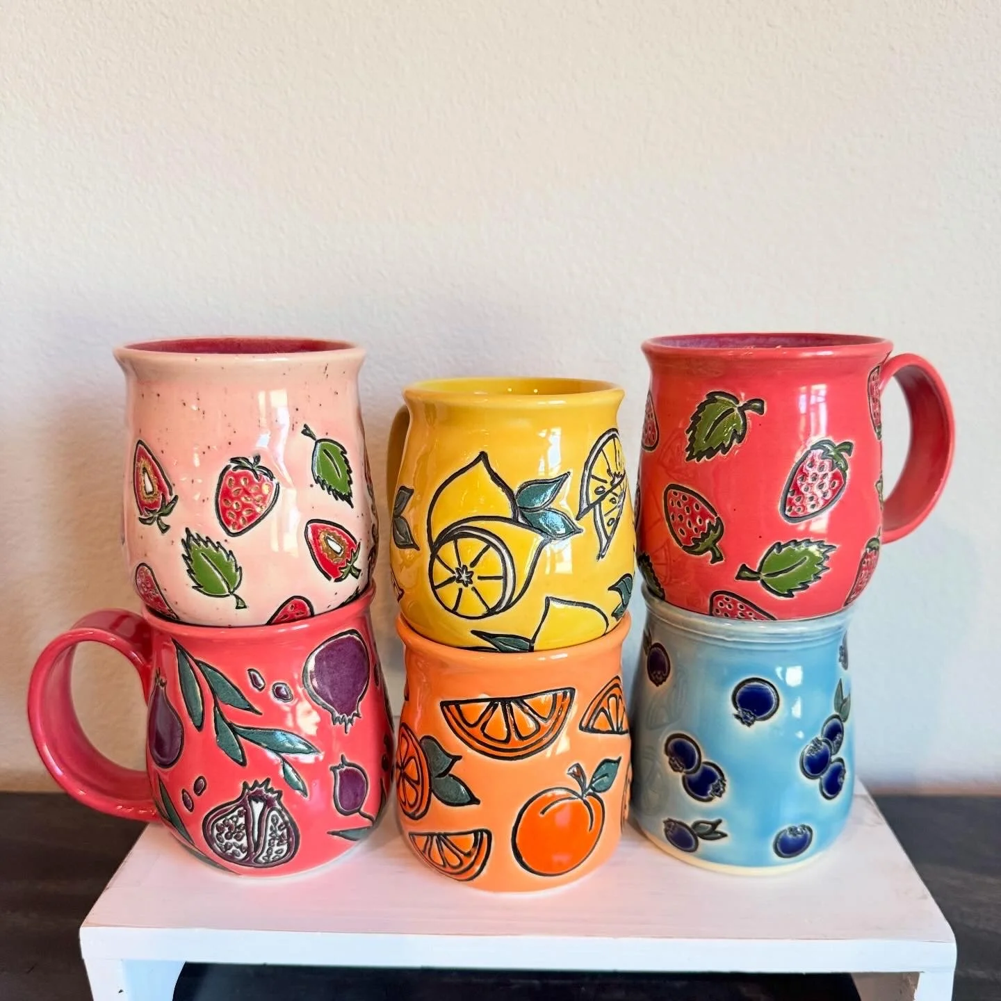 Six colorful ceramic mugs with fruit patterns placed on a white stand against a plain wall.