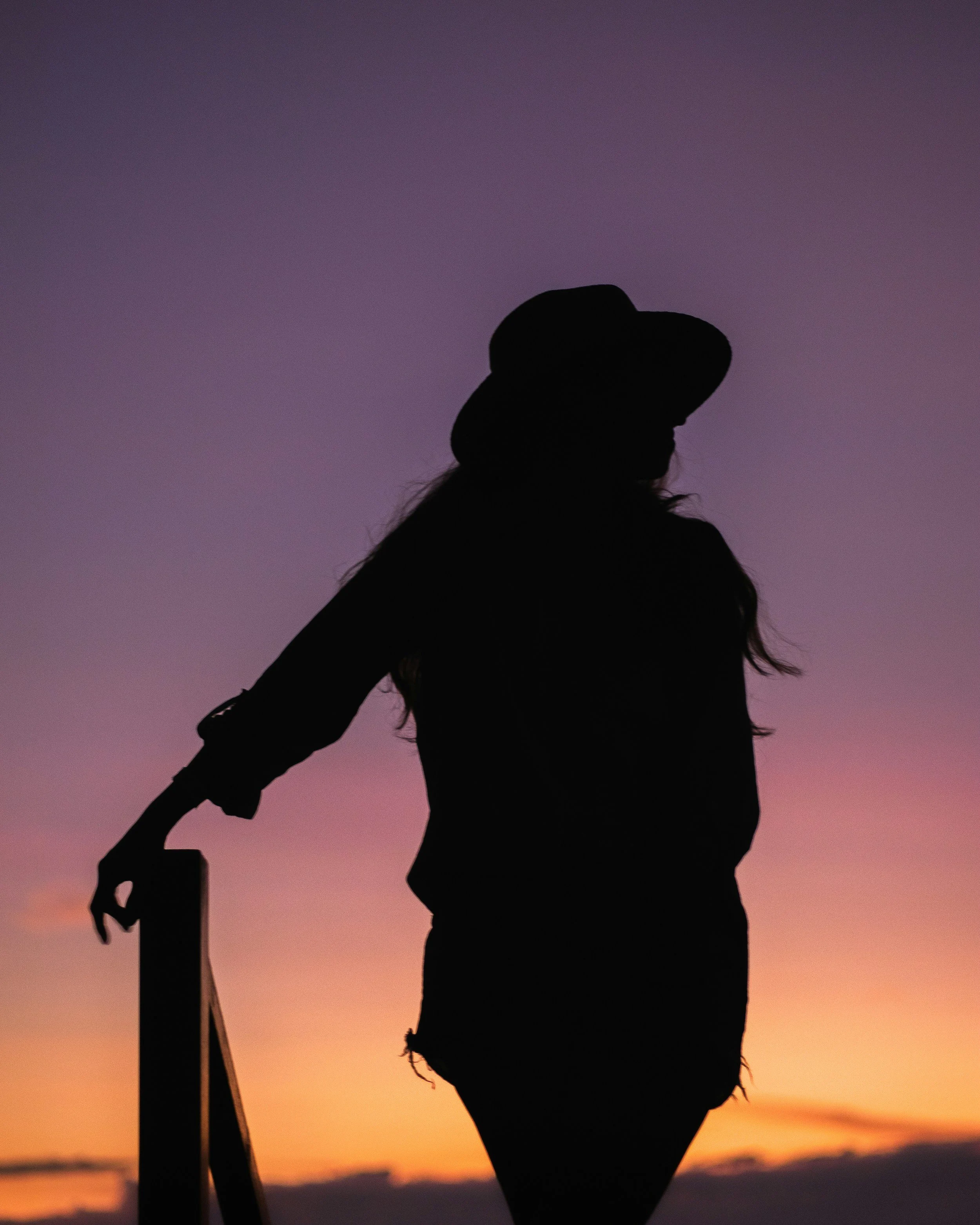 Silhouette of a person wearing a hat against a sunset sky with purple and orange hues.