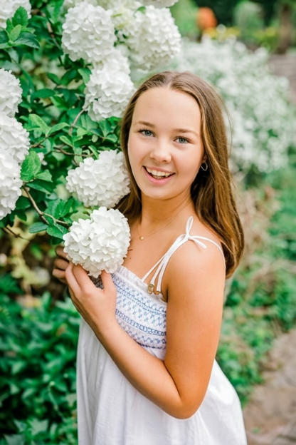 A young girl with long brown hair smiling while holding a white hydrangea flower next to a bush of hydrangeas.