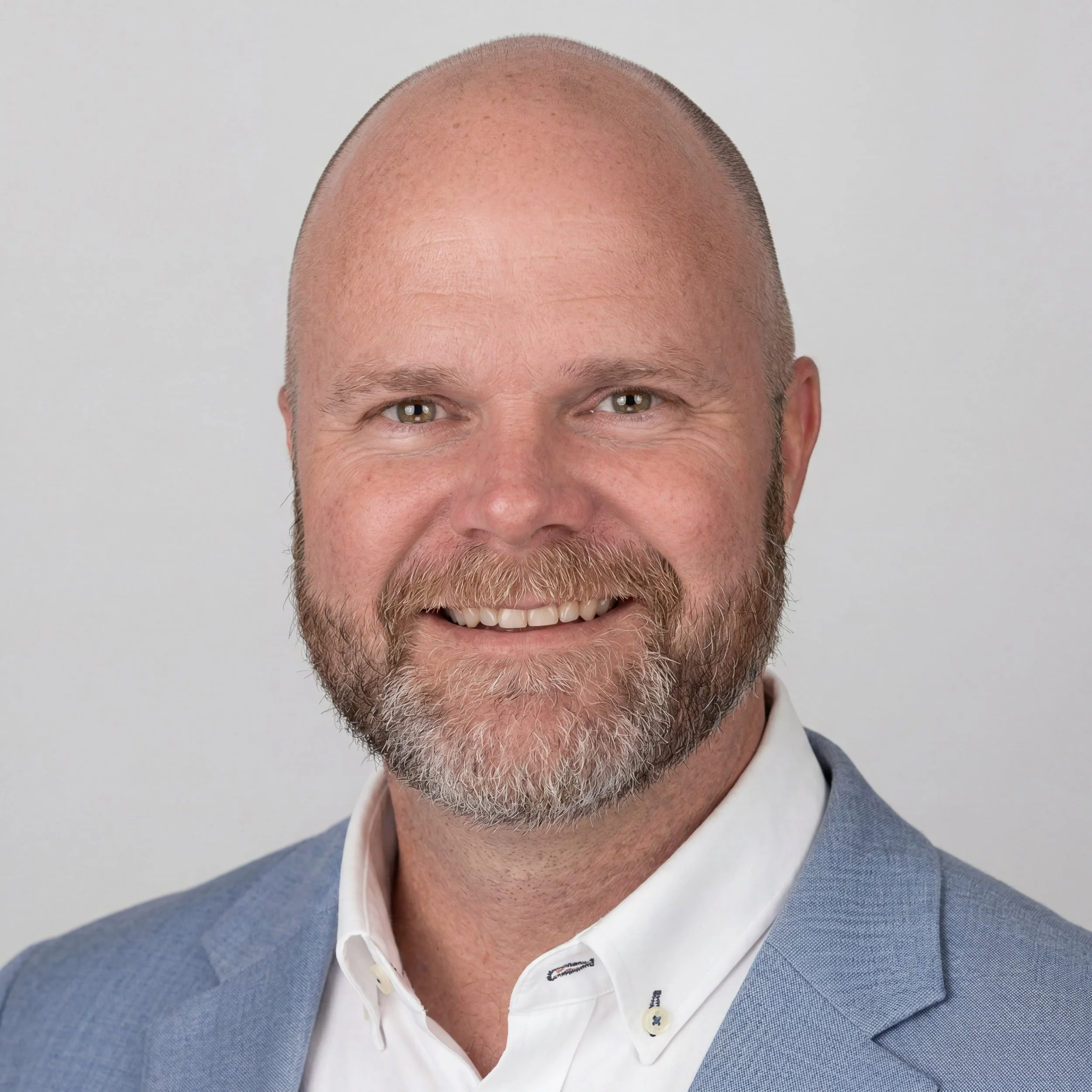 Close-up of a smiling man with a shaved head, white beard, wearing a white shirt and a light blue blazer.