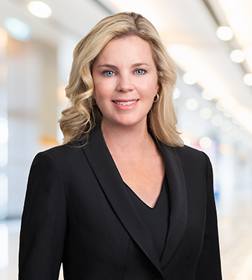 A woman with blonde hair wearing a black blazer, smiling, in an indoor setting with bright lighting.
