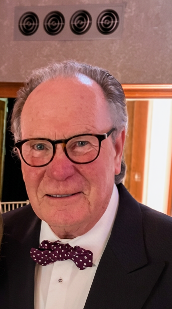 A man with glasses and a bow tie in formal attire, indoors with a doorway and electronic vents in the background.