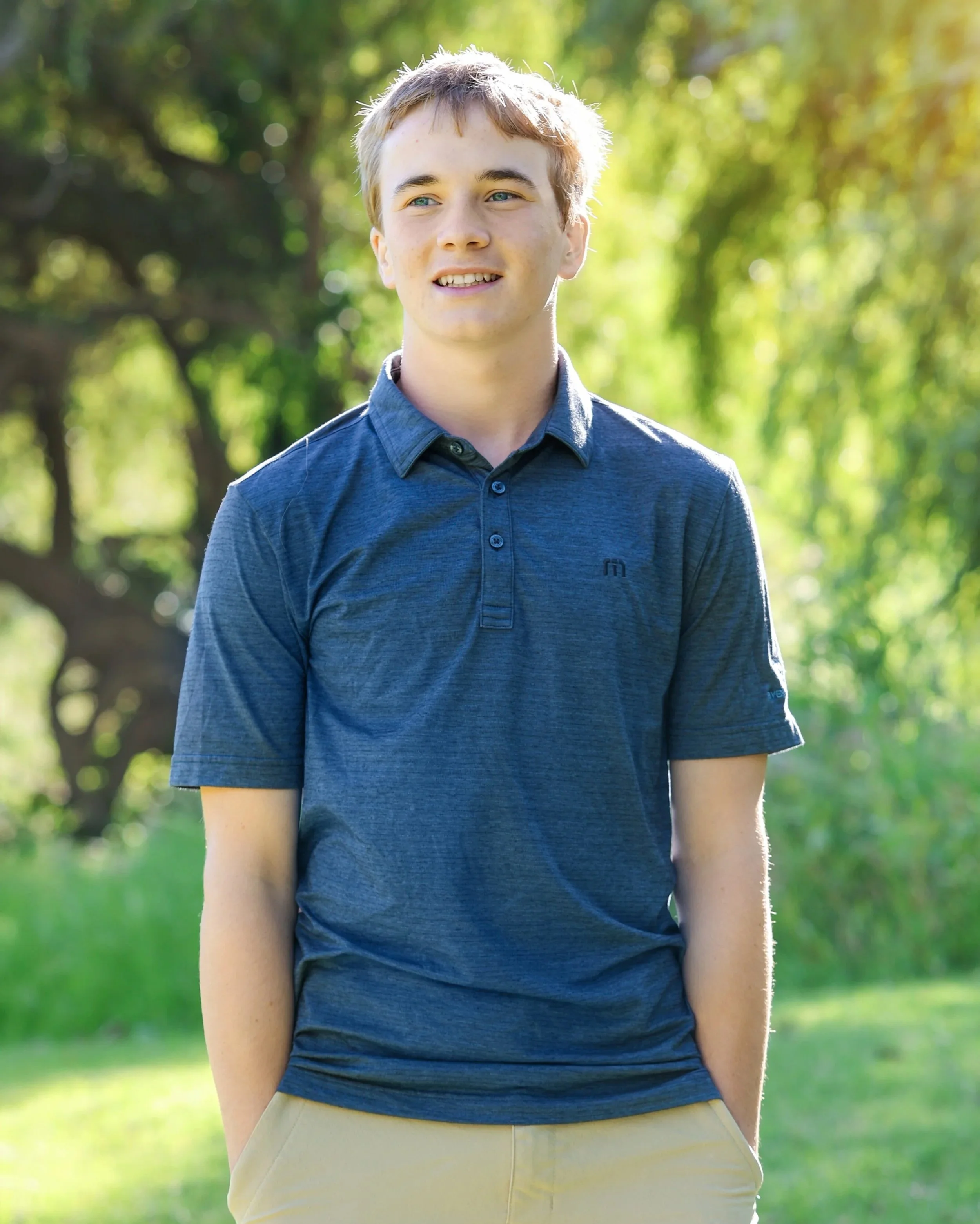 Portrait of a young man standing outdoors in a park with green trees in the background, smiling and looking slightly to the side. He has light brown hair, blue eyes, and is wearing a blue collared shirt and beige pants.