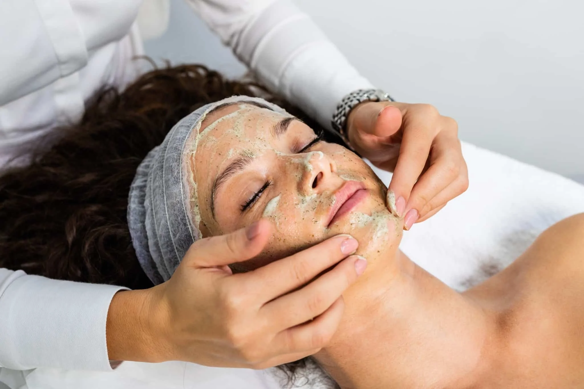 A woman lying down with her eyes closed, receiving a facial treatment in a spa setting, with a skincare mask on her face and a beautician gently touching her cheek.