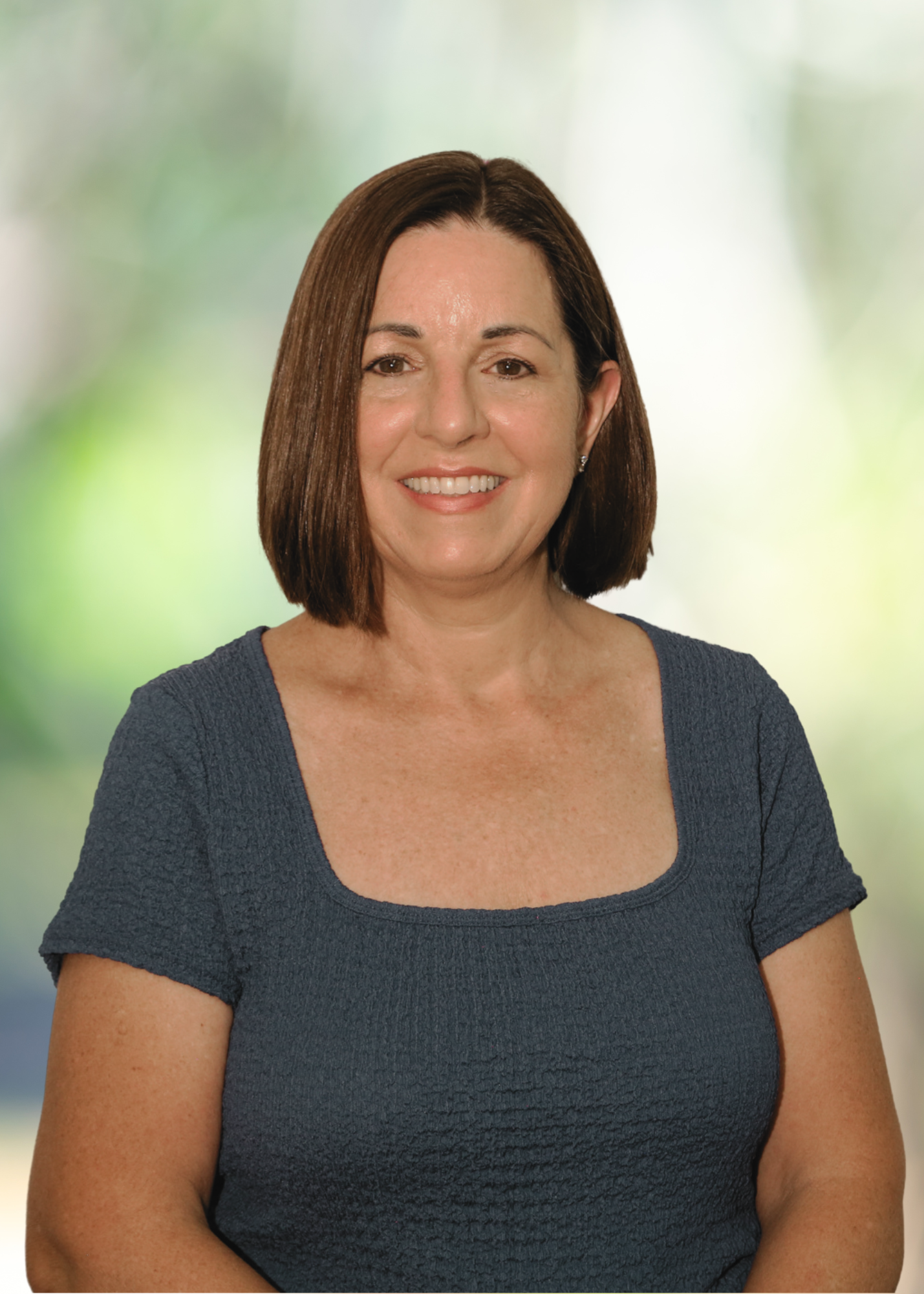 A woman with shoulder-length brown hair wearing a dark gray textured short-sleeve top, smiling in front of a blurred green background.