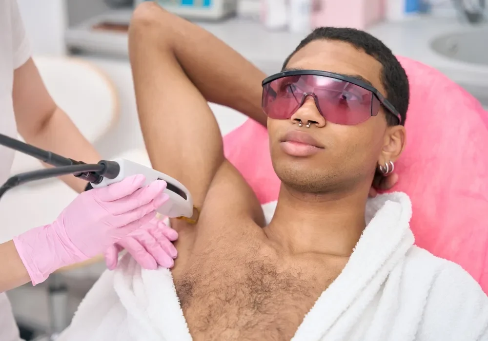 A man with dark hair, wearing pink-tinted safety goggles, earrings, and a septum pierce, is lying back on a pink pillow and getting laser hair removal on his armpit from a technician wearing pink gloves.