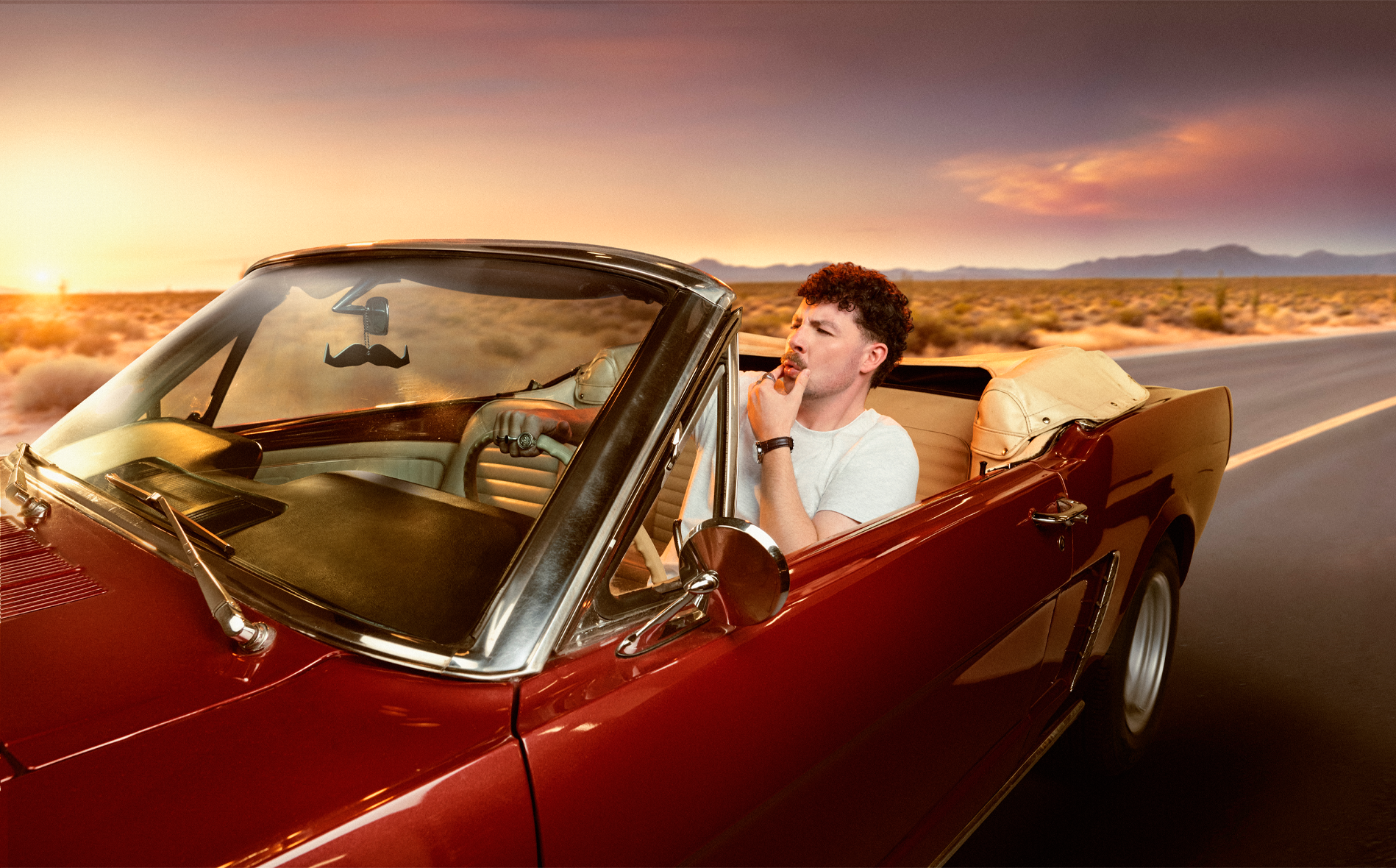 jack james ryan sits in a red vintage convertible car on a desert road at sunset, thinking with a finger on his lips.