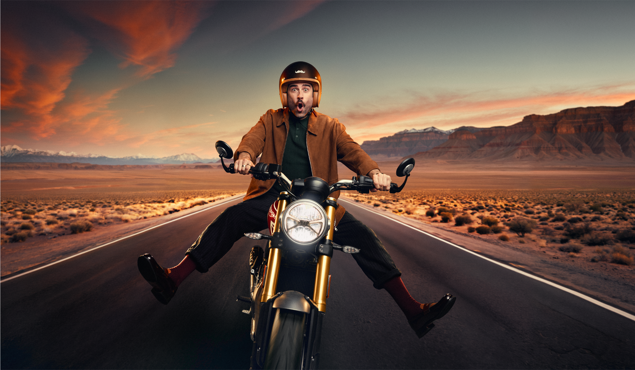 Matt Johnson in a helmet is riding a motorcycle on an open road in a desert landscape during sunset, with mountains in the background and a colorful sky.