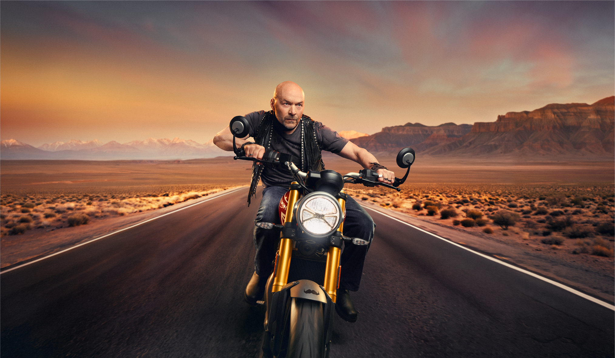 Simon Rimmer riding a motorcycle on an empty desert highway at sunset, with mountains in the background.