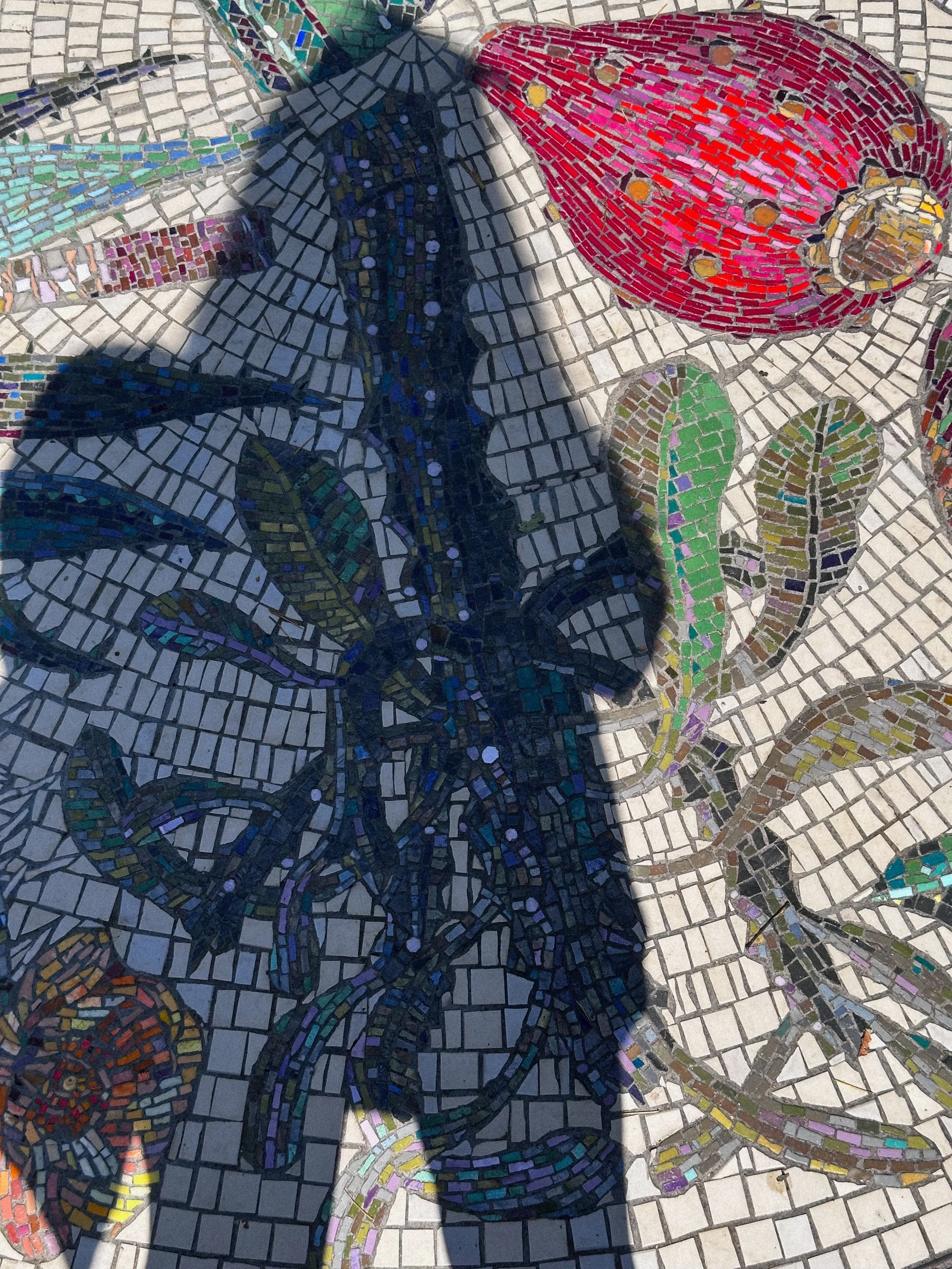 A colorful mosaic tile artwork featuring a butterfly, a ladybug, and an abstract plant scene, partially shadowed on a tiled surface.