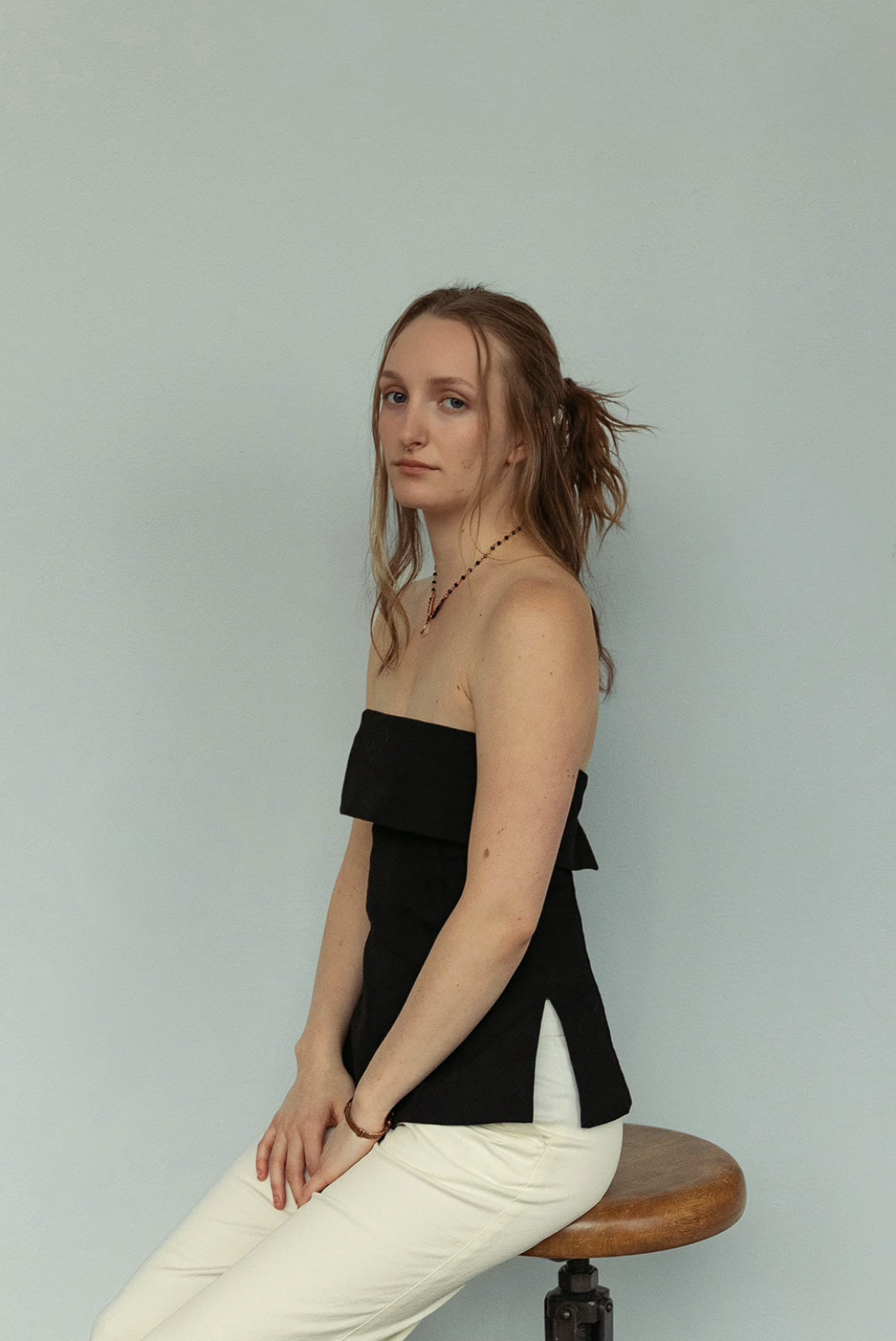 A young woman with straight and pinned up blonde hair sitting on a wooden stool against a plain light-colored wall, wearing a black strapless top and white pants.