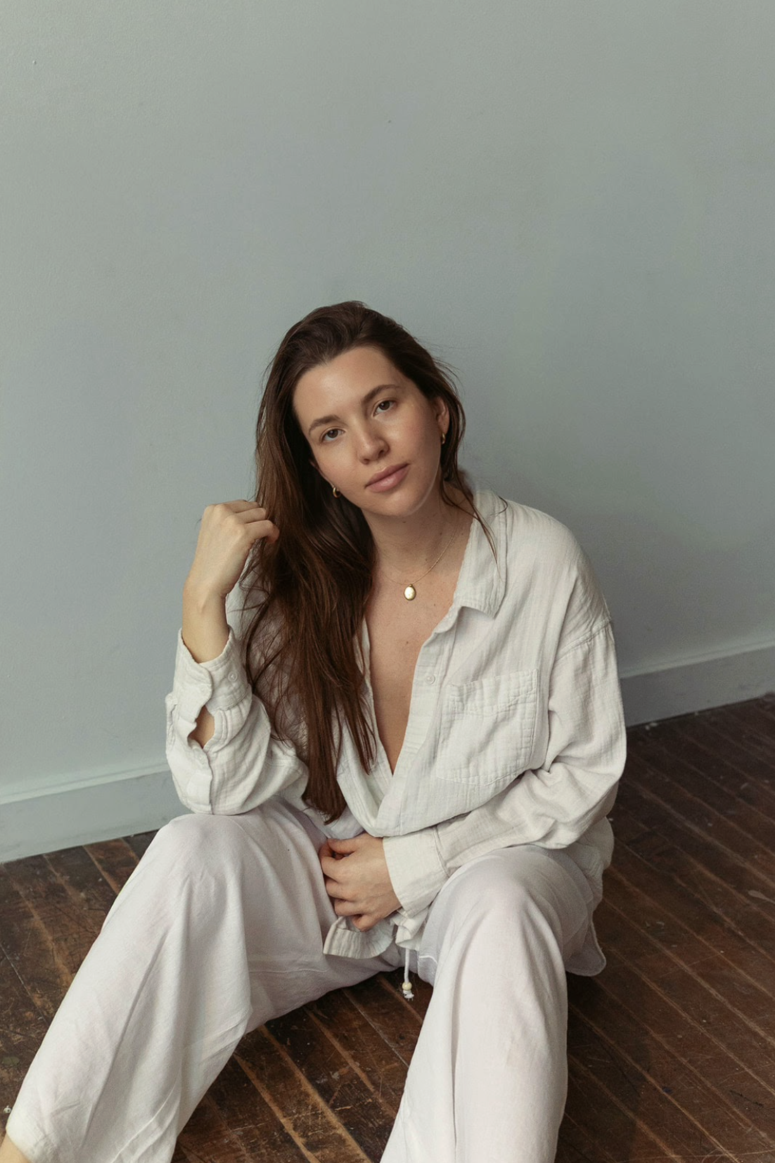 A woman with long brown hair, wearing a beige button-up shirt and beige pants, sitting on a wooden floor near a plain wall, looking at the camera with a relaxed expression.