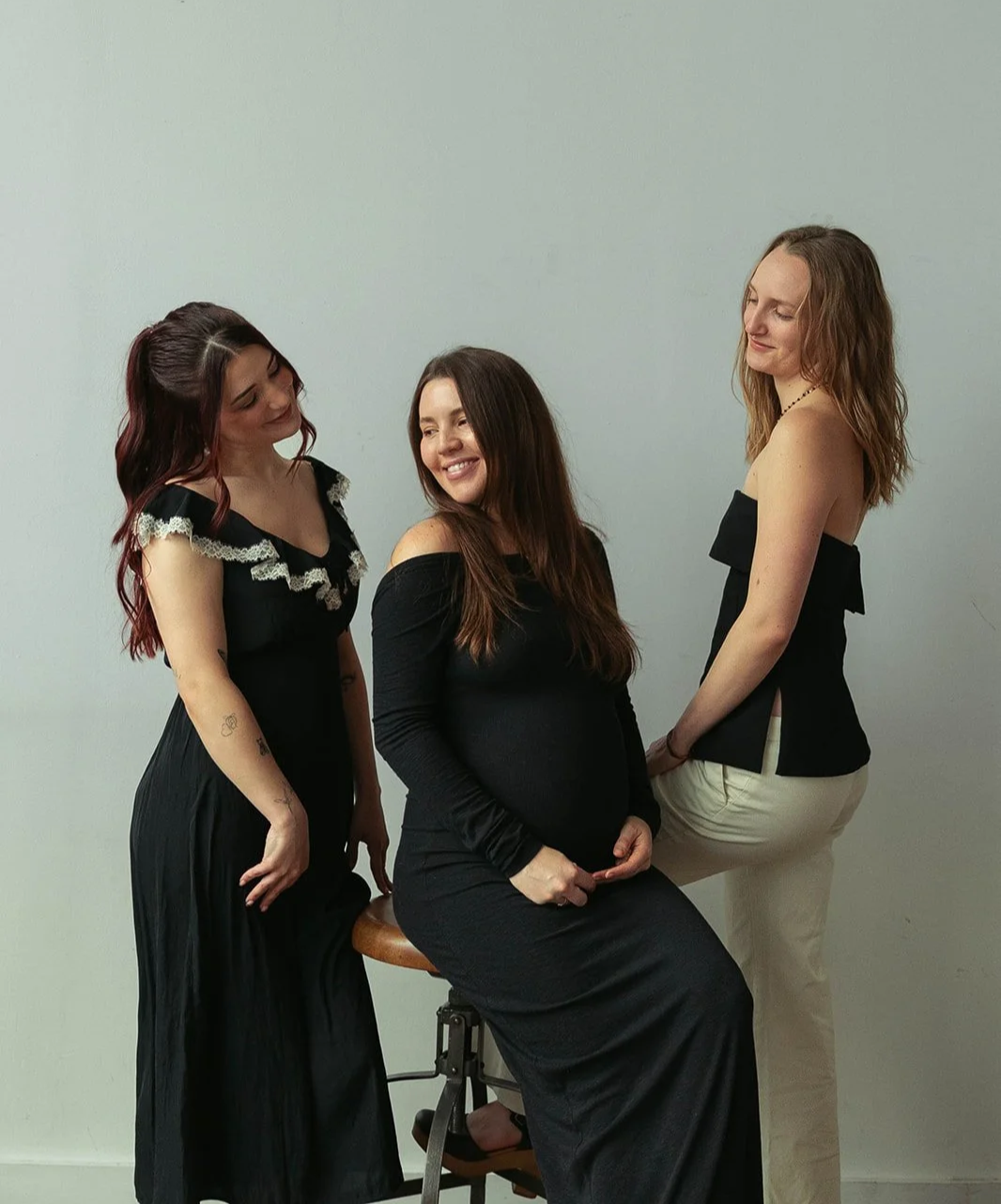 Three women wearing black and white clothing in a photo studio. One woman is sitting on a stool, the other two standing beside her, all looking at each other and smiling.