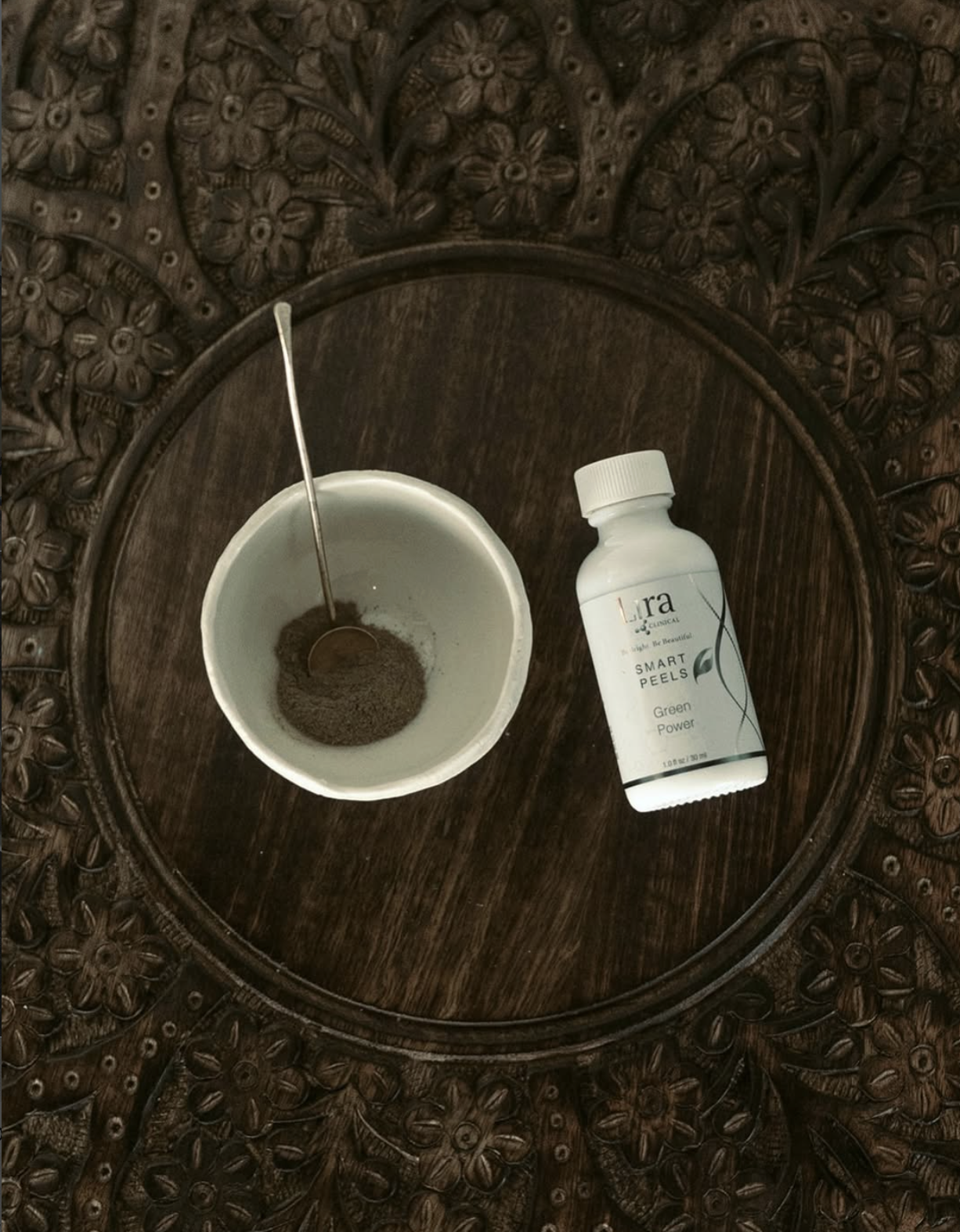 A small bowl with powdered skin supplement, a spoon inside, and a bottle labeled 'Smart Peels' on a wooden tray with decorative carved edges.