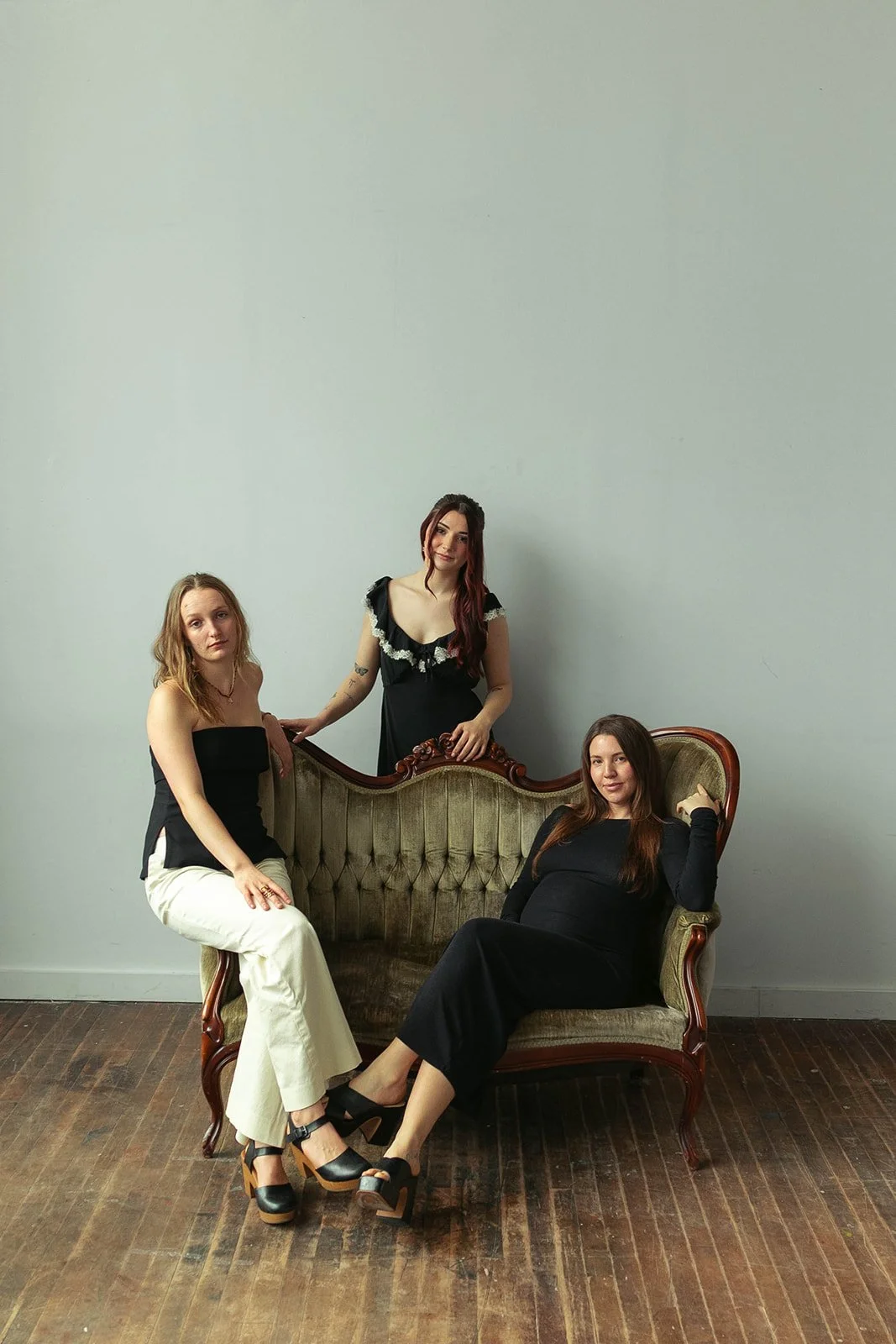 Three women in black outfits posing around a vintage green upholstered sofa in a studio with a plain light-colored wall and wooden floor.