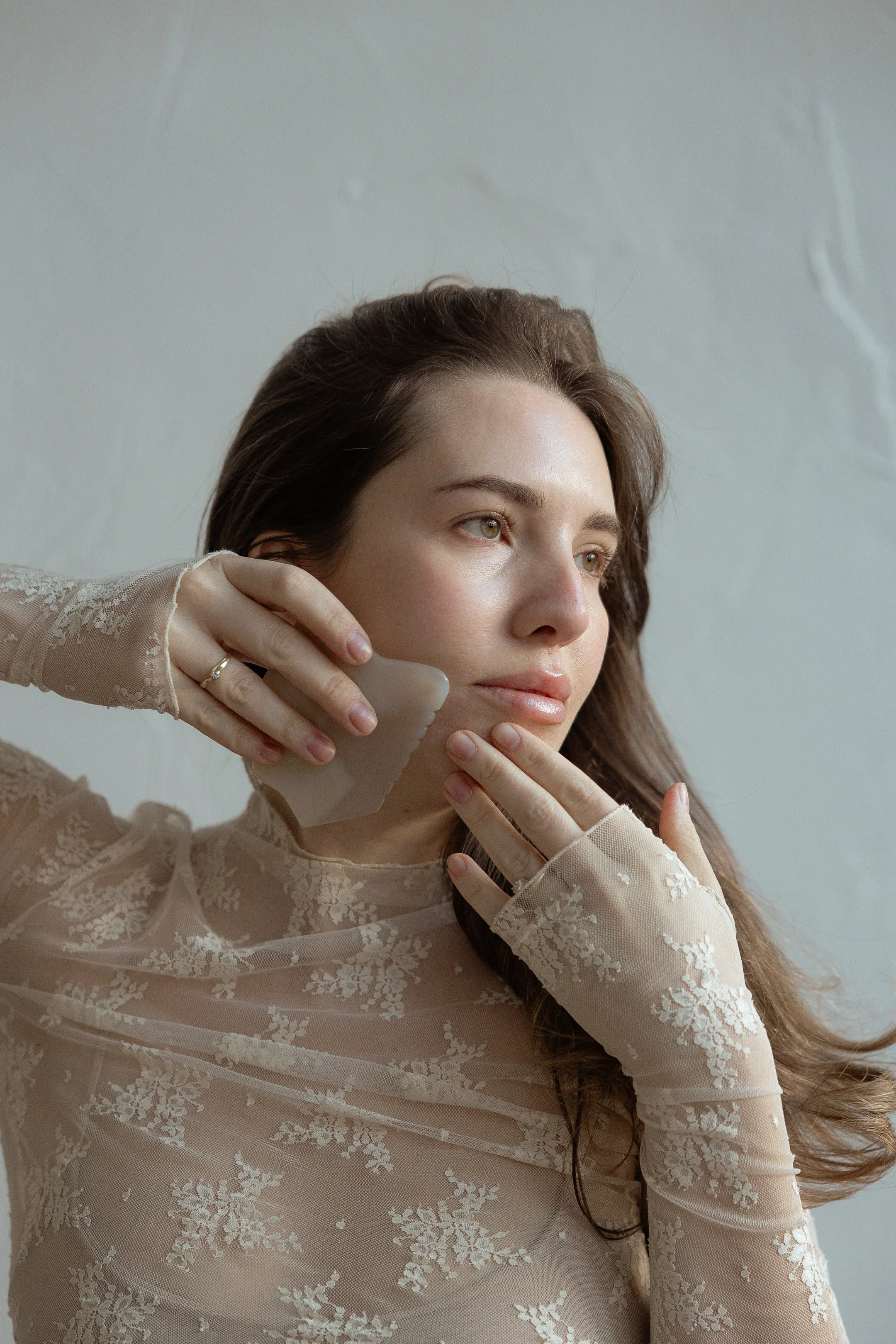 A woman with long brown hair in a lace beige top is using a gua sha stone on her cheek, looking thoughtful and serene.