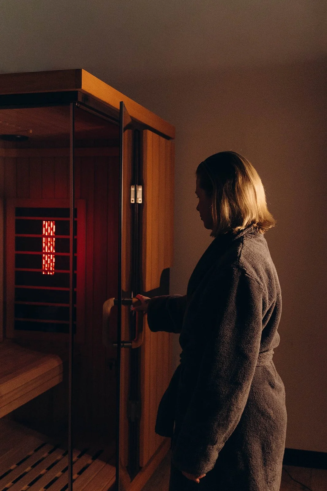 A woman in a bathrobe standing outside of a sauna, holding the door open, and ready to walk in, with the sauna's interior red light visible.