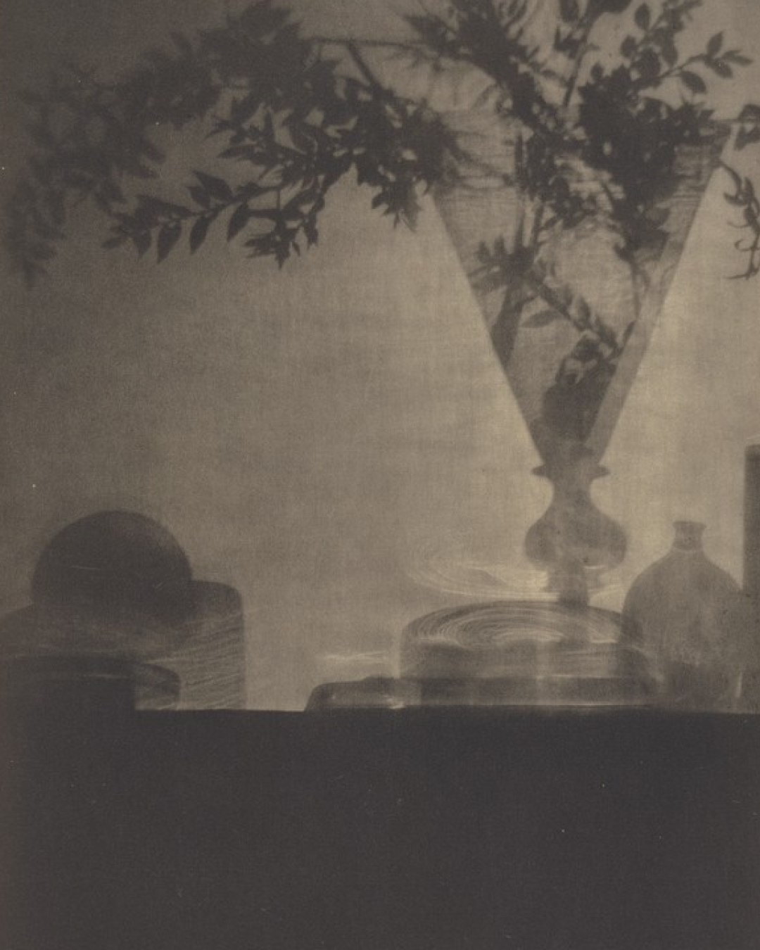 Shadow of a vase with plant on a wall, with objects and containers on a table.