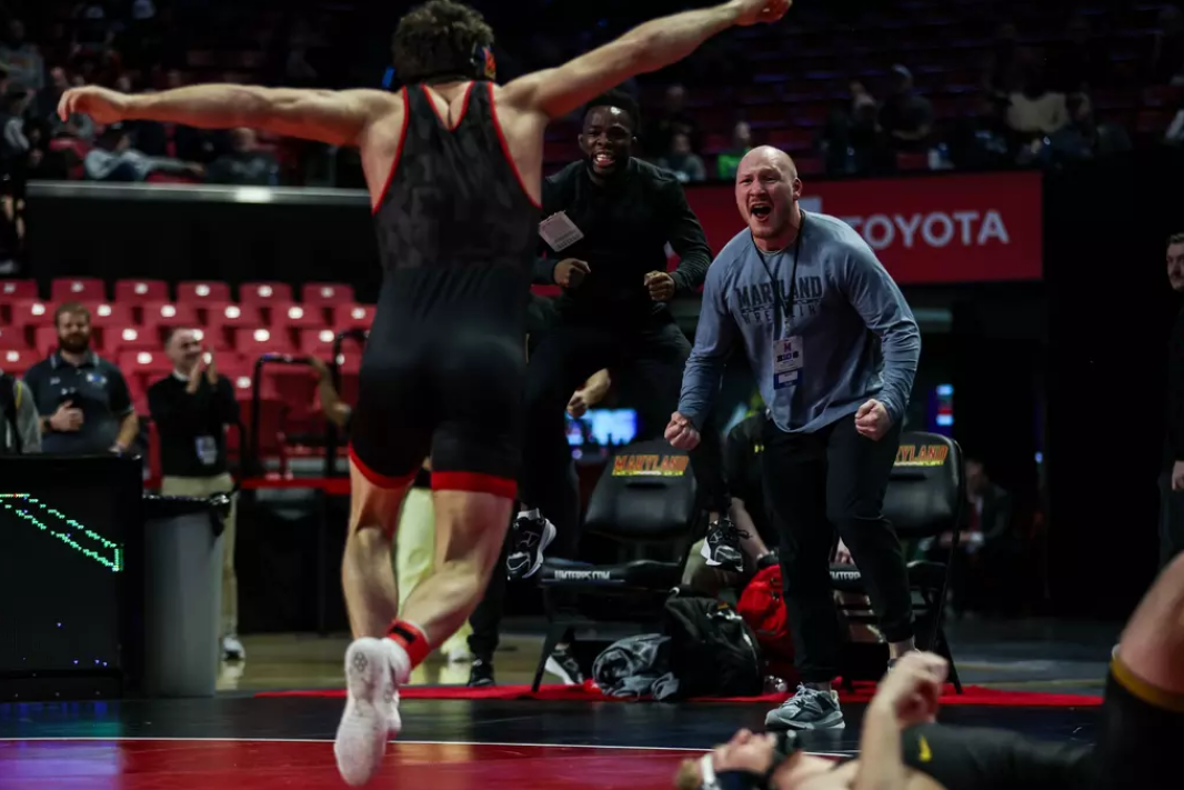 A wrestler celebrates a victory with outstretched arms while a teammate and coach cheer excitedly in the background.