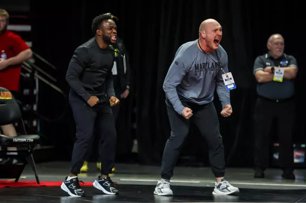 Two wrestlers are celebrating passionately on the mat, with fists clenched and mouths open, during a wrestling match at an event, while spectators and officials look on in the background.