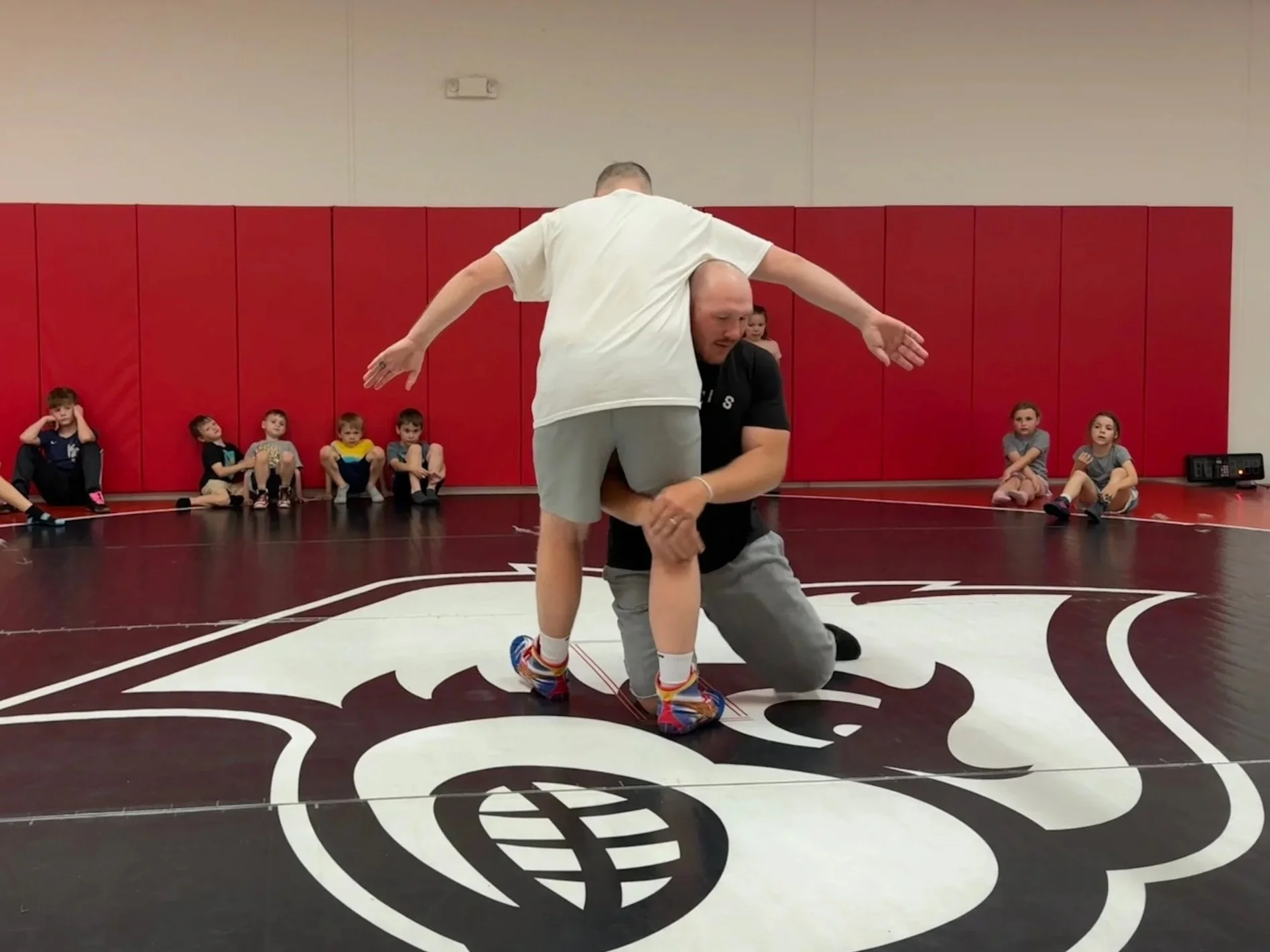 Two men demonstrate wrestling techniques on a gymnasium floor with kids seated along the wall observing.